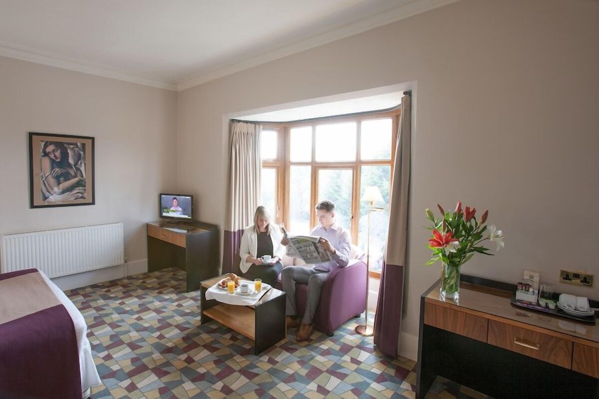 Ein Hotelzimmer mit Bett, Schreibtisch, Fernseher, Fenster mit Vorhängen, einem Couchtisch mit Frühstück und einer Vase mit Blumen.