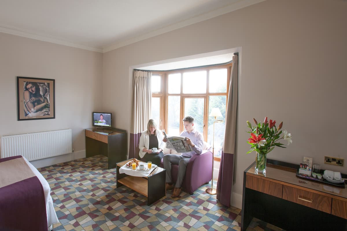 A hotel room with a bed, a desk with a computer, a coffee table with breakfast, a window with curtains, and a painting on the wall.