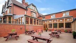 A courtyard with multiple wooden picnic tables and potted plants, surrounded by a brick building with large windows and a balcony. - Staindrop Hotel (Sheffield)