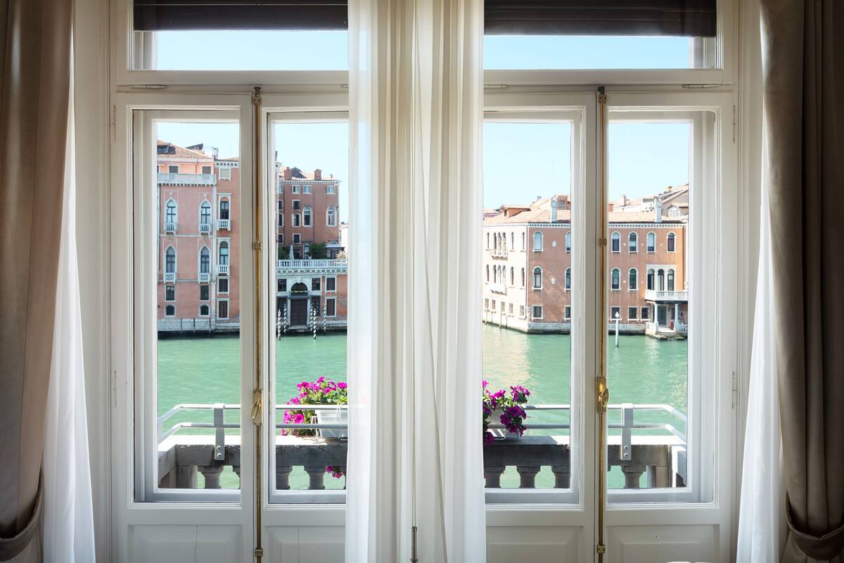 A view from a window with white curtains, overlooking a canal with buildings and a bridge.