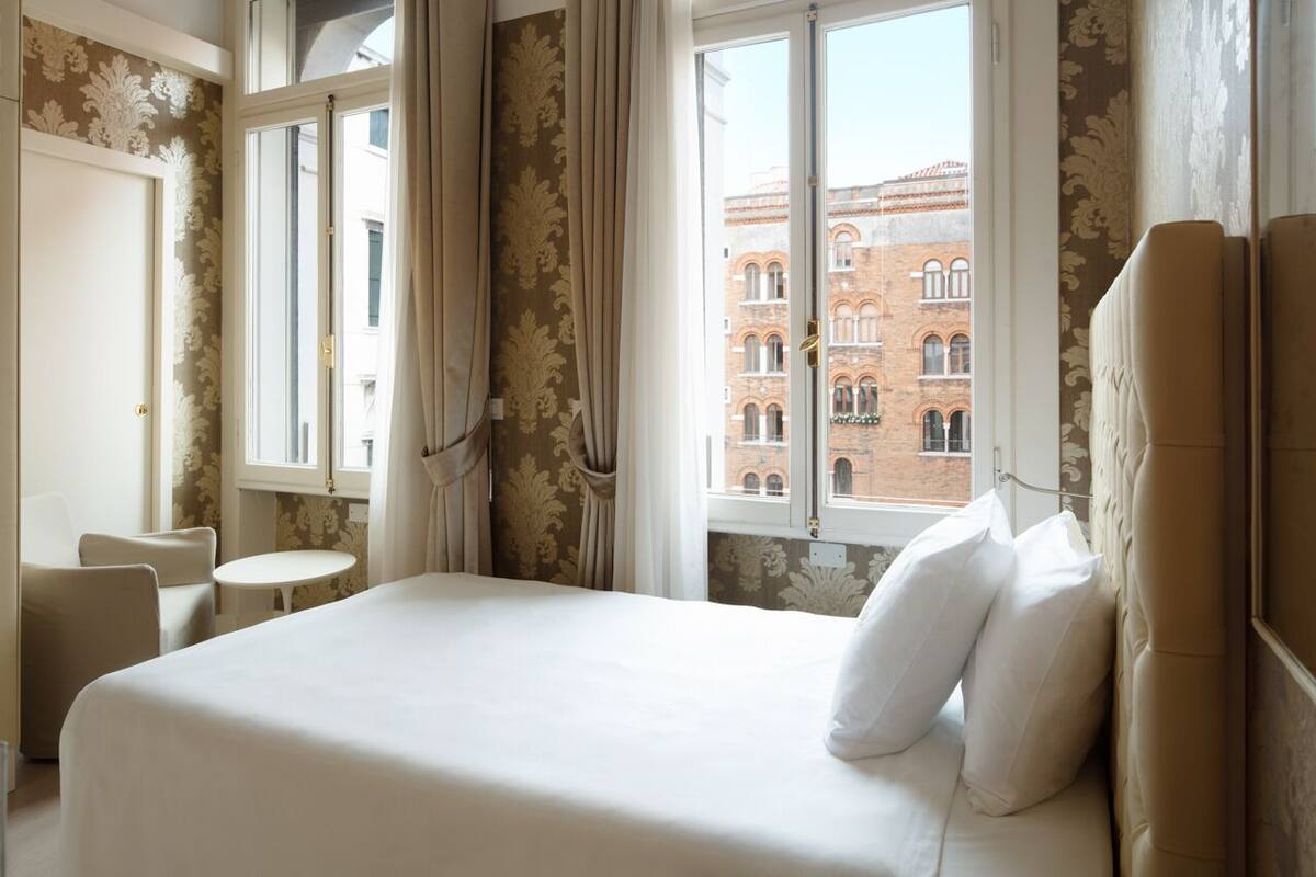 A hotel room with a large bed, two chairs, a small table, and a view of a historic building through the window.