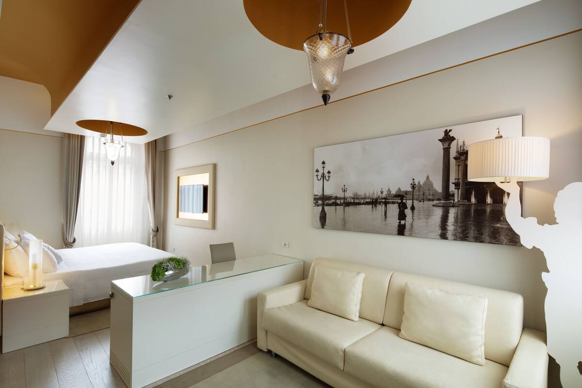 A modern hotel room with a bed, a glass desk, a sofa, a large cityscape painting, and a chandelier.