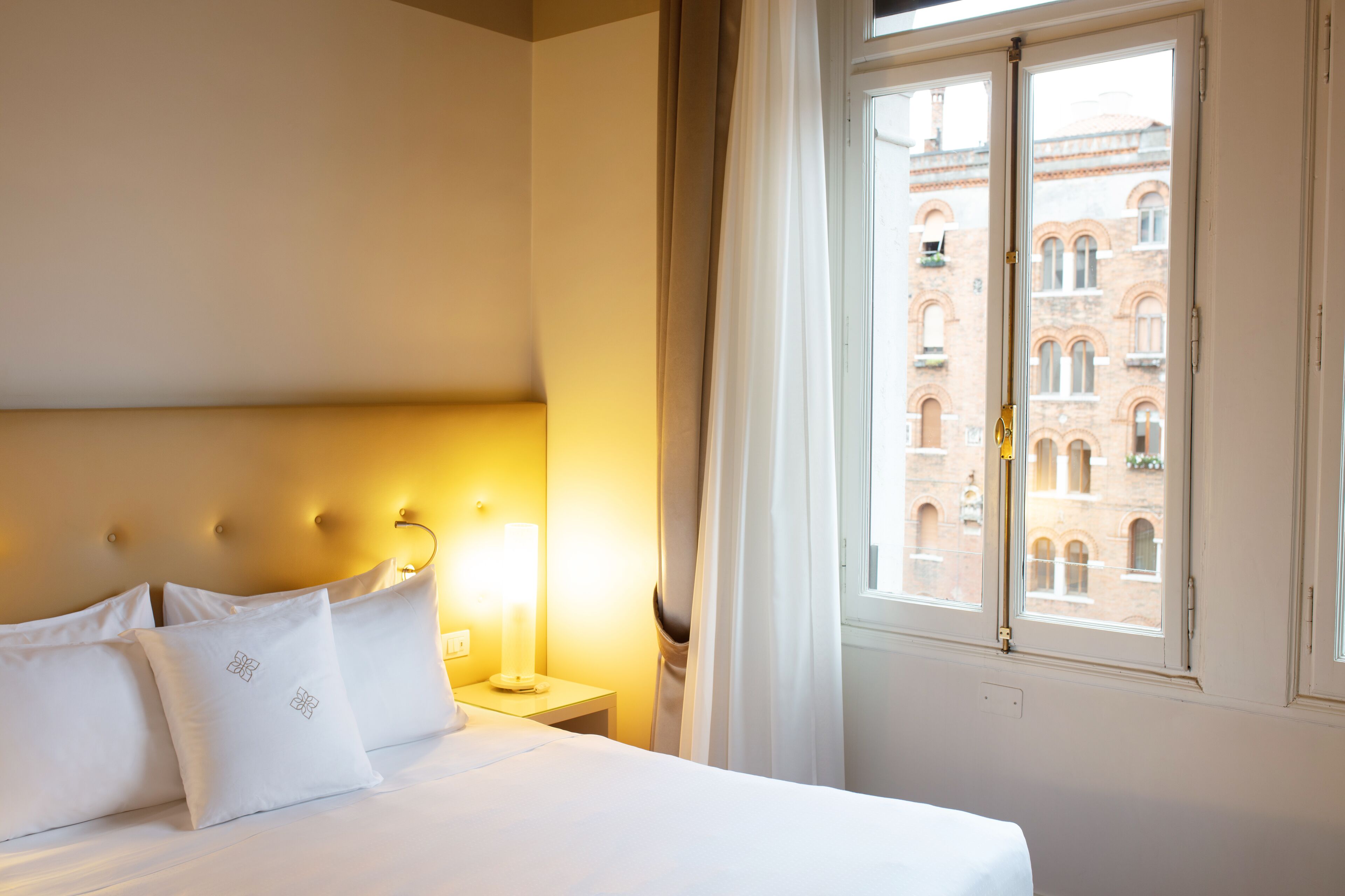 A hotel room with a bed, a nightstand, a lamp, and a window with a view of a building.
