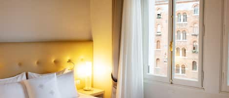 A hotel room with a bed, a nightstand, a lamp, and a window with a view of a building.
