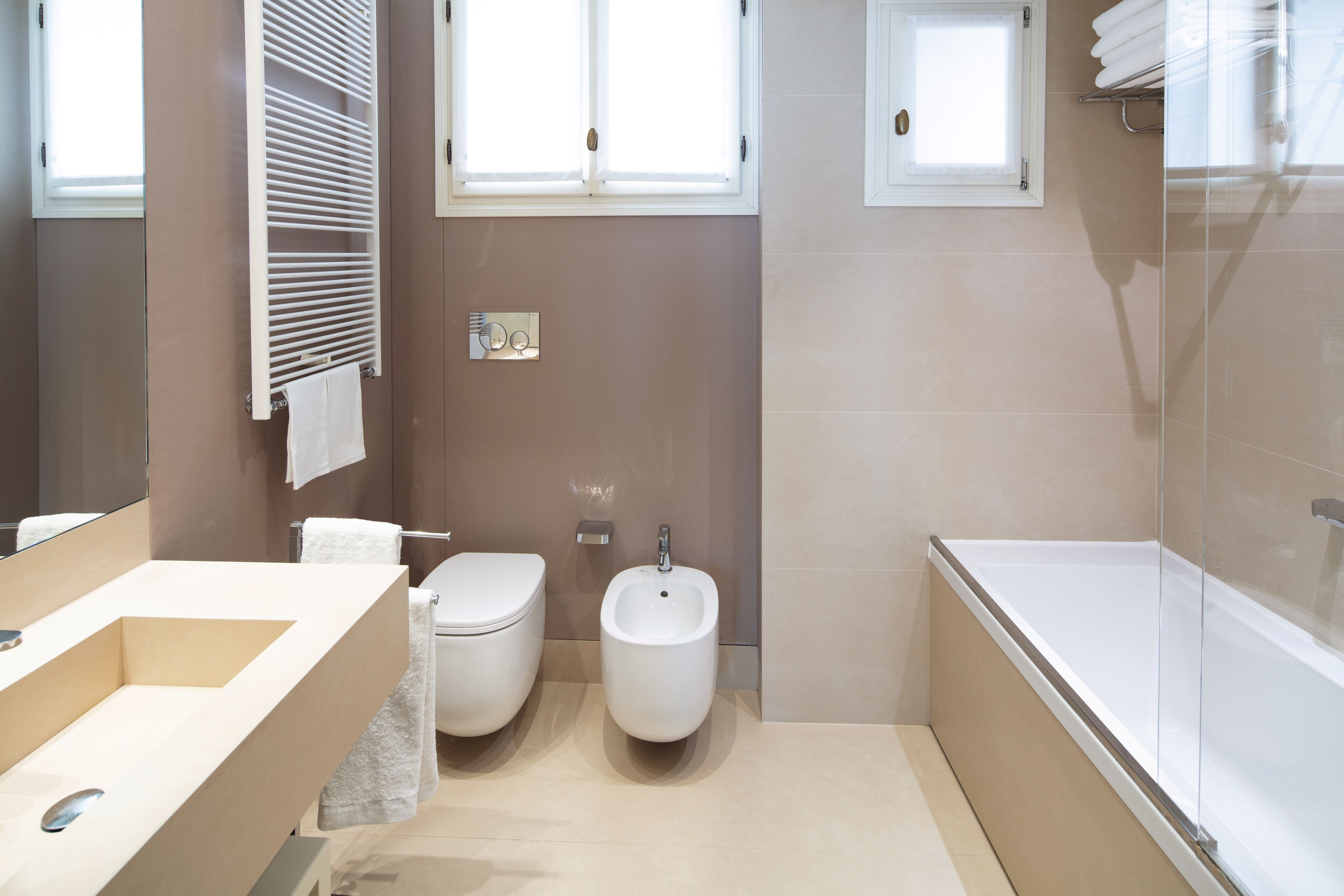 A modern bathroom with a beige tiled floor, a white sink, a toilet, a bidet, and a bathtub. There are two windows providing natural light.