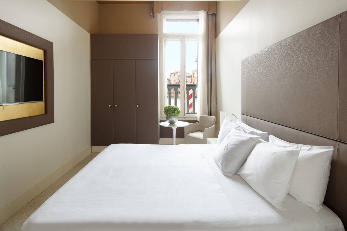 A hotel room with a large bed, a TV mounted on the wall, a small round table, and a view of the city through a window.