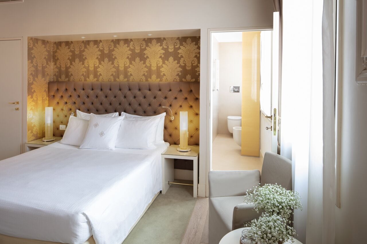 A hotel room with a large bed, a nightstand with a lamp, a bedside table with a vase of flowers, and a view of the bathroom through an open door.