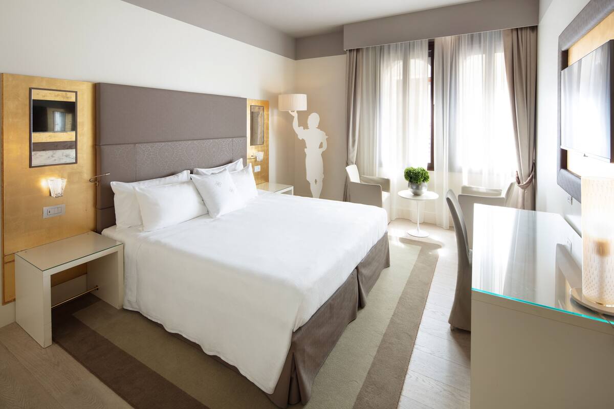 A hotel room with a large bed, a desk, a chair, a TV, and a small table with a plant.