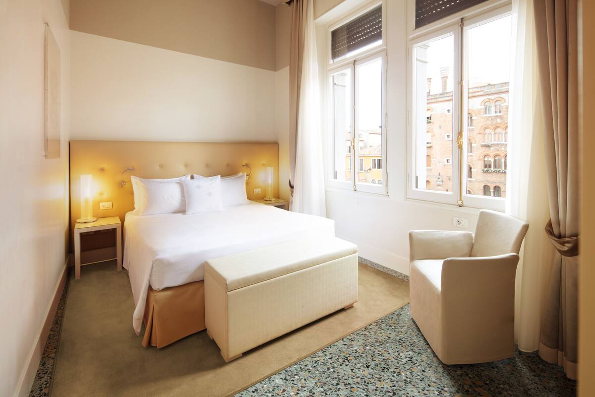 A hotel room with a large bed, bedside lamps, a chair, and a view of buildings through the window.