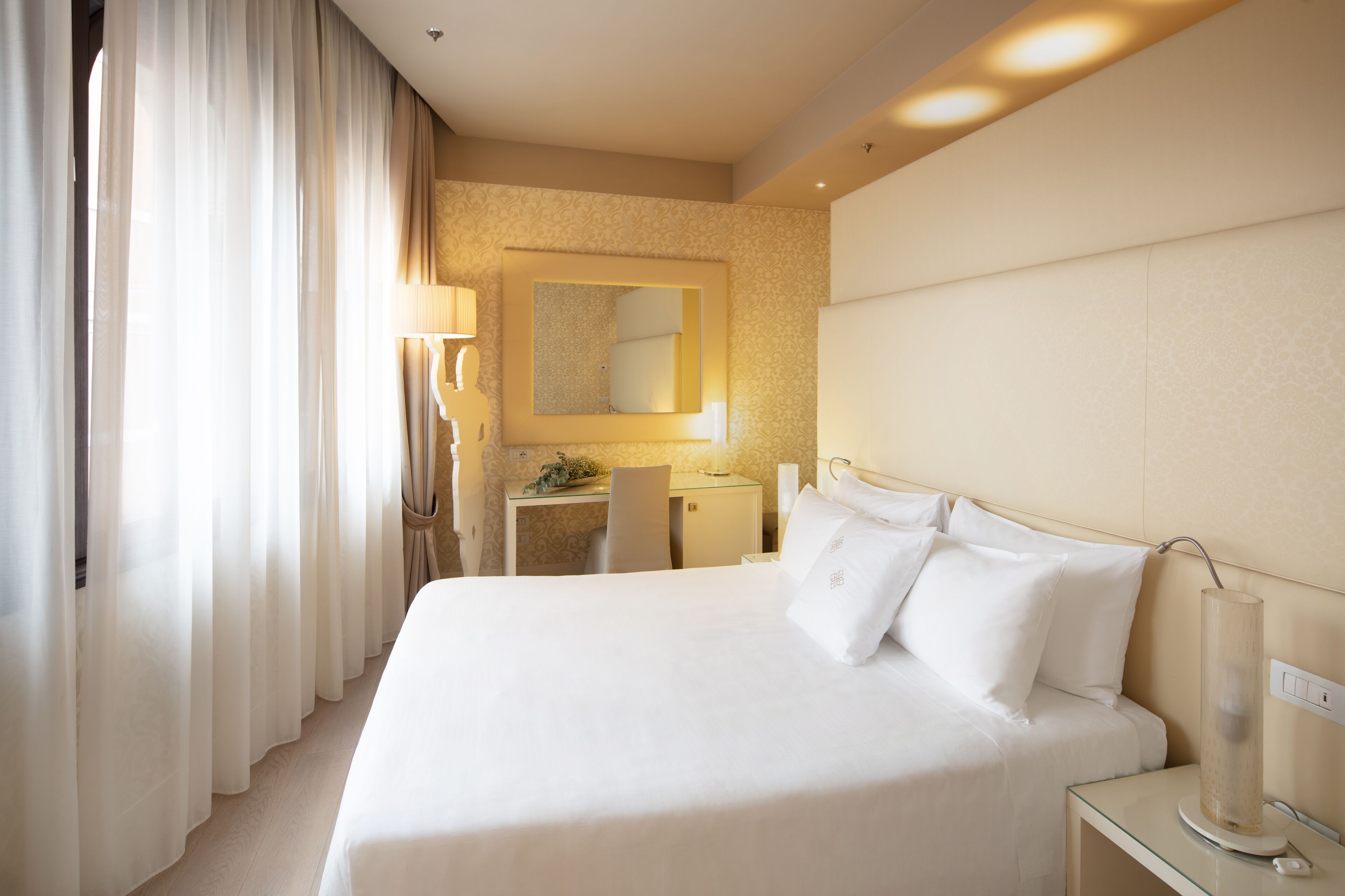 A hotel room with a large bed, a desk, a chair, a lamp, and a mirror.
