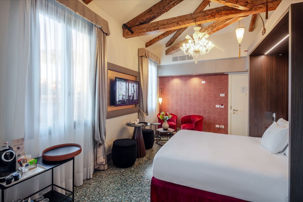 A hotel room with a large bed, a TV mounted on the wall, a red armchair, and a small table with a vase of flowers.
