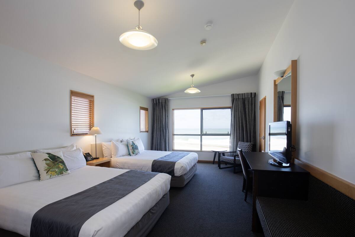 Ein Hotelzimmer mit zwei Betten, einem Schreibtisch mit Fernseher, einem Sessel, einer Lampe und Blick auf den Strand.