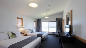 Une chambre d’hôtel avec deux lits, un bureau avec une télévision, une chaise, une lampe et une vue sur la plage.