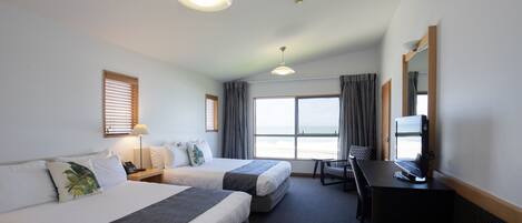 Habitación de hotel con dos camas, un escritorio con televisor, una silla, una lámpara y vista a la playa.