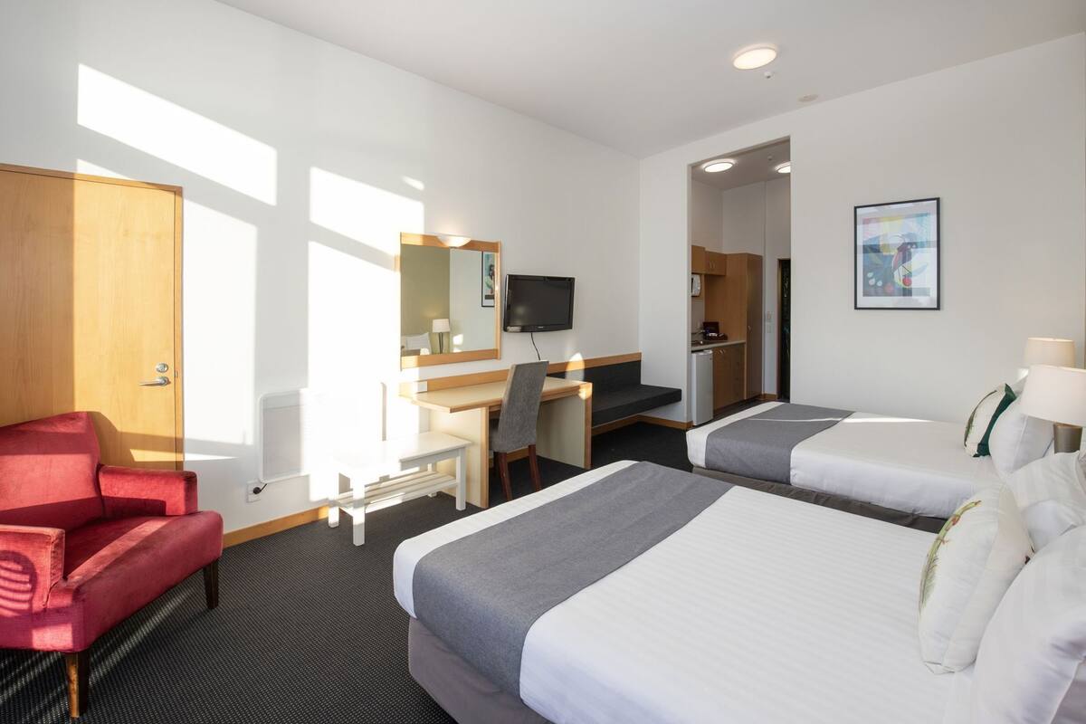 Ein Hotelzimmer mit zwei Betten, einem Schreibtisch, einem Stuhl, einem Fernseher und einer kleinen Küchenzeile.