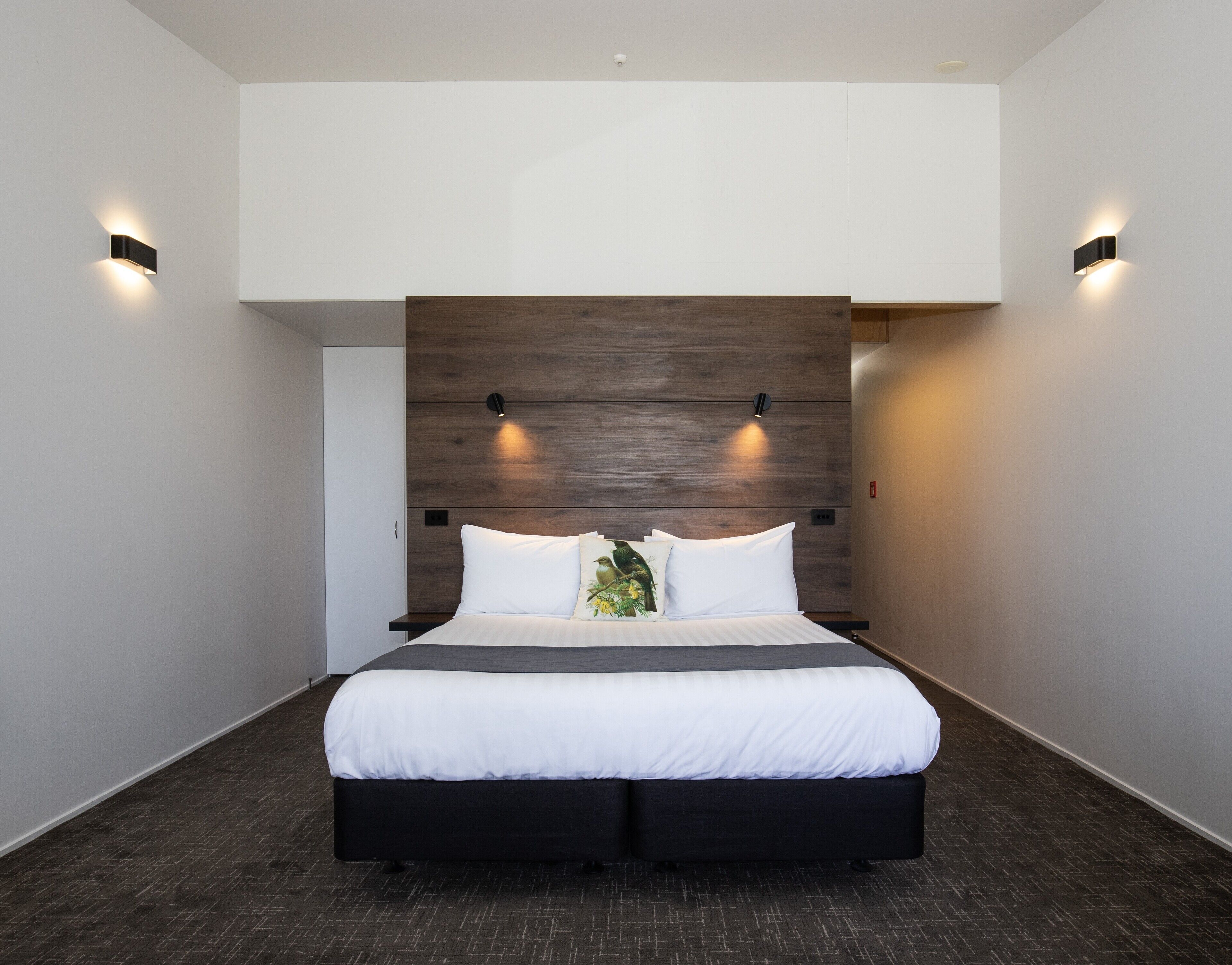 A modern hotel room with a large bed, wooden headboard, and two wall-mounted lights.
