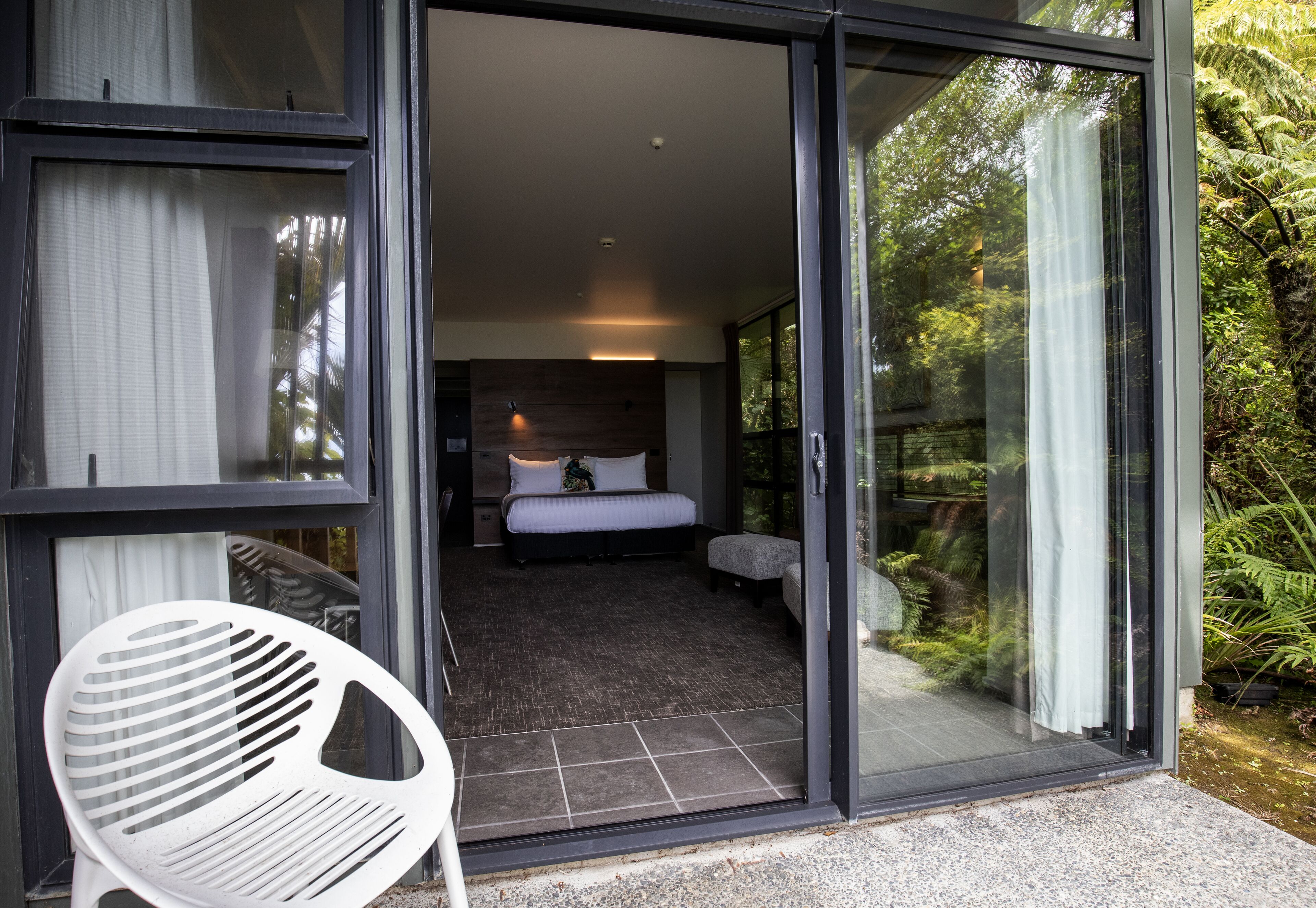 A modern bedroom with a large bed, a small seating area, and a glass door leading to a garden.