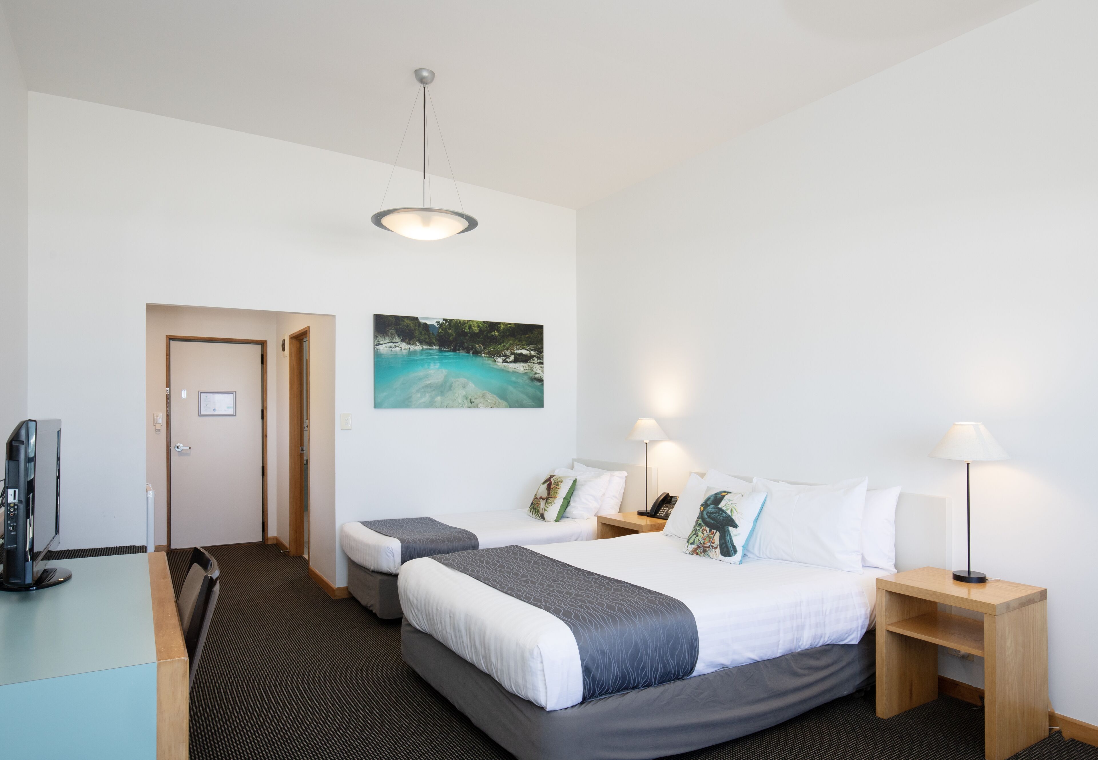 Habitación de hotel con dos camas, un escritorio, un televisor, un cuadro de playa y una lámpara.