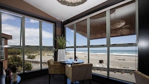 Una habitación con grandes ventanales que dan a una playa, una mesa con sillas y una planta en maceta.