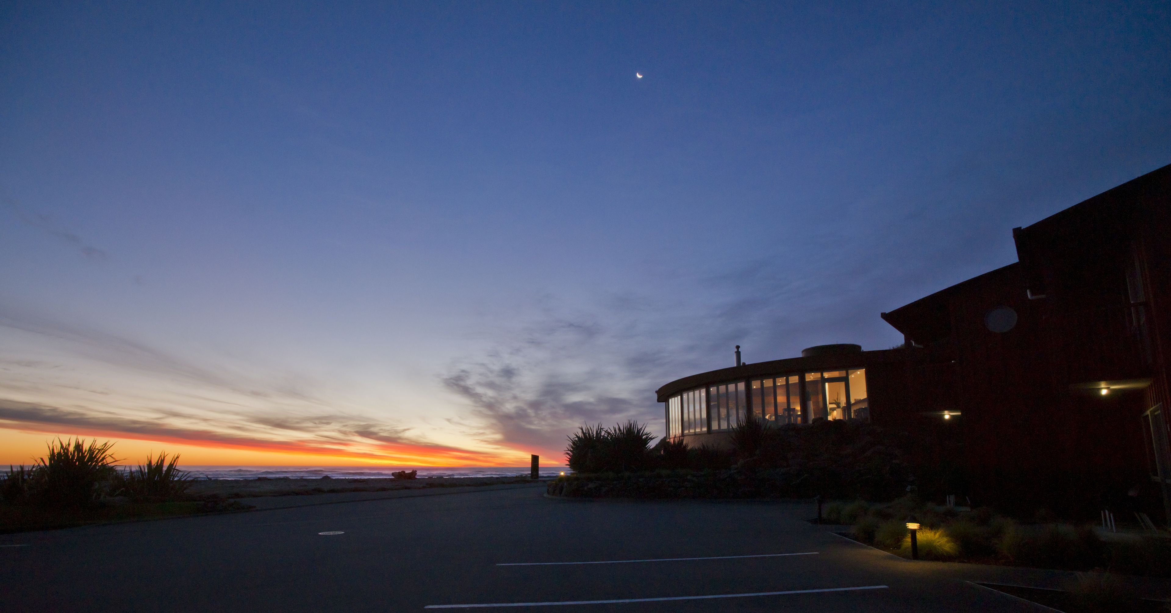 A building with a curved facade and large windows, situated next to a parking lot with a view of the sunset over the ocean.