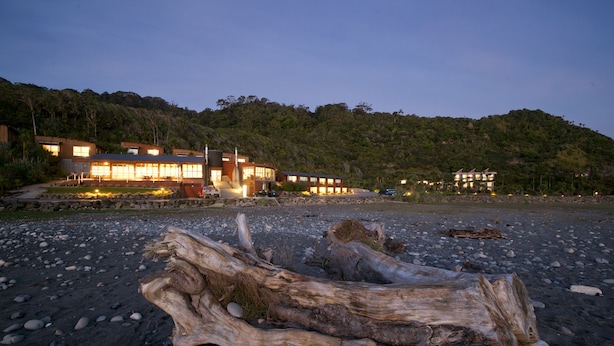 Un edificio moderno con ventanas amplias y un camino de piedra que conduce a él, ubicado en una playa de guijarros con ramas flotantes al frente.