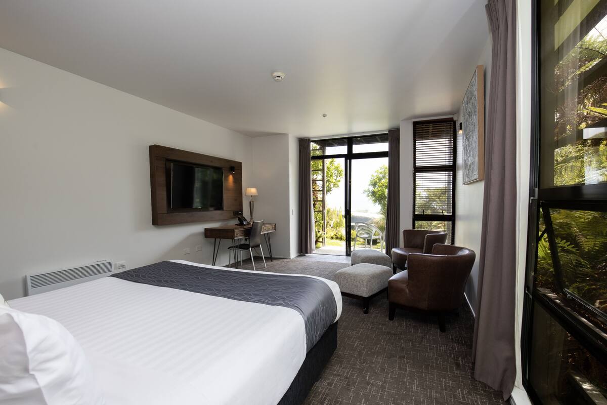 Ein Hotelzimmer mit einem großen Bett, einem Schreibtisch mit Stuhl, einer kleinen Couch und einem großen Fenster mit Blick auf Bäume.