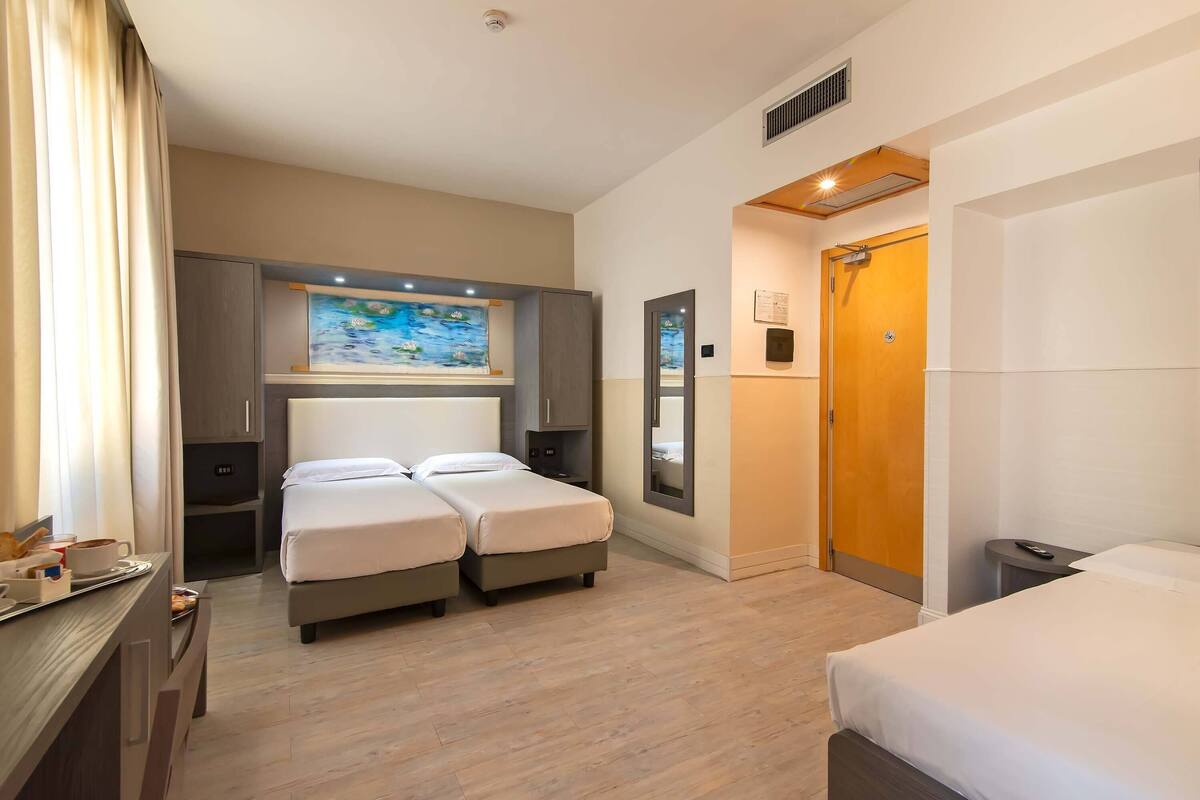 Una camera d'albergo con un letto matrimoniale, una scrivania con una sedia, un tavolino con un vassoio contenente articoli per la colazione, un quadro appeso e una porta in legno.