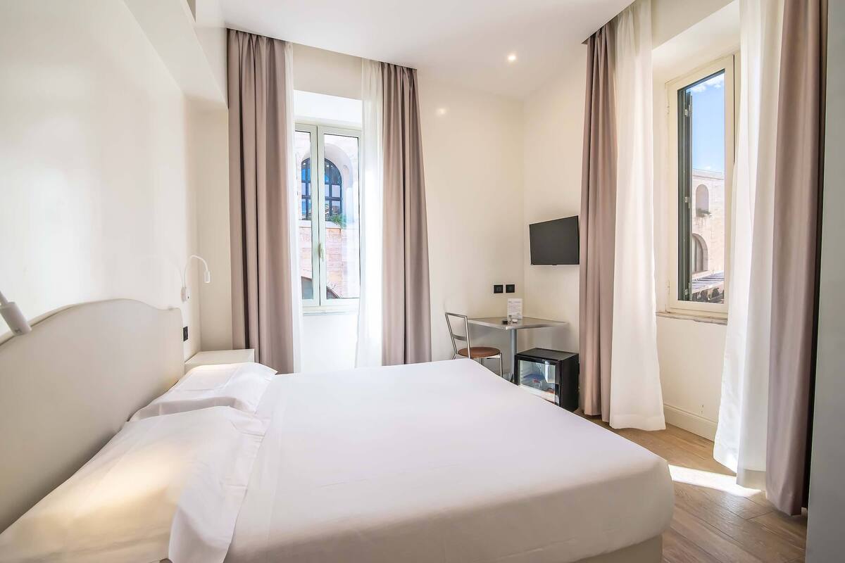 Una camera d'albergo con un letto, una scrivania, una televisione e una finestra con tende.