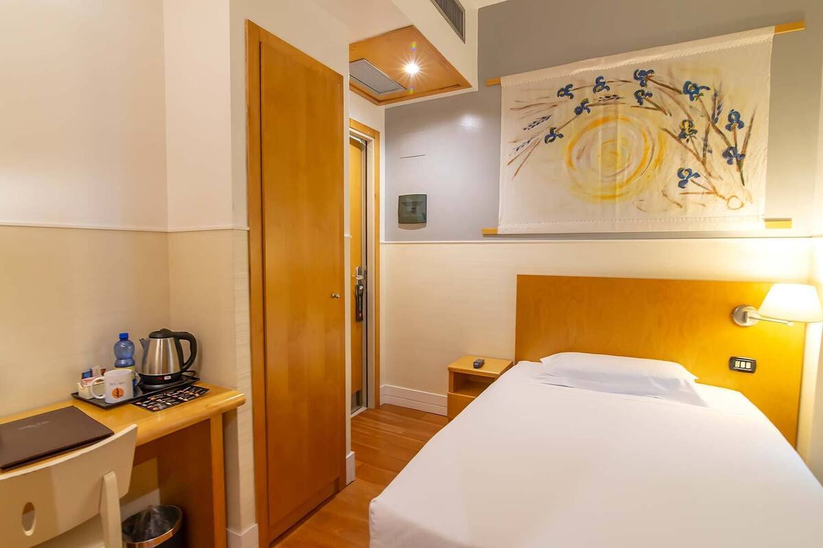 Una camera d'albergo con un letto, una scrivania con bollitore, una bottiglia d'acqua e una macchina del caffè.