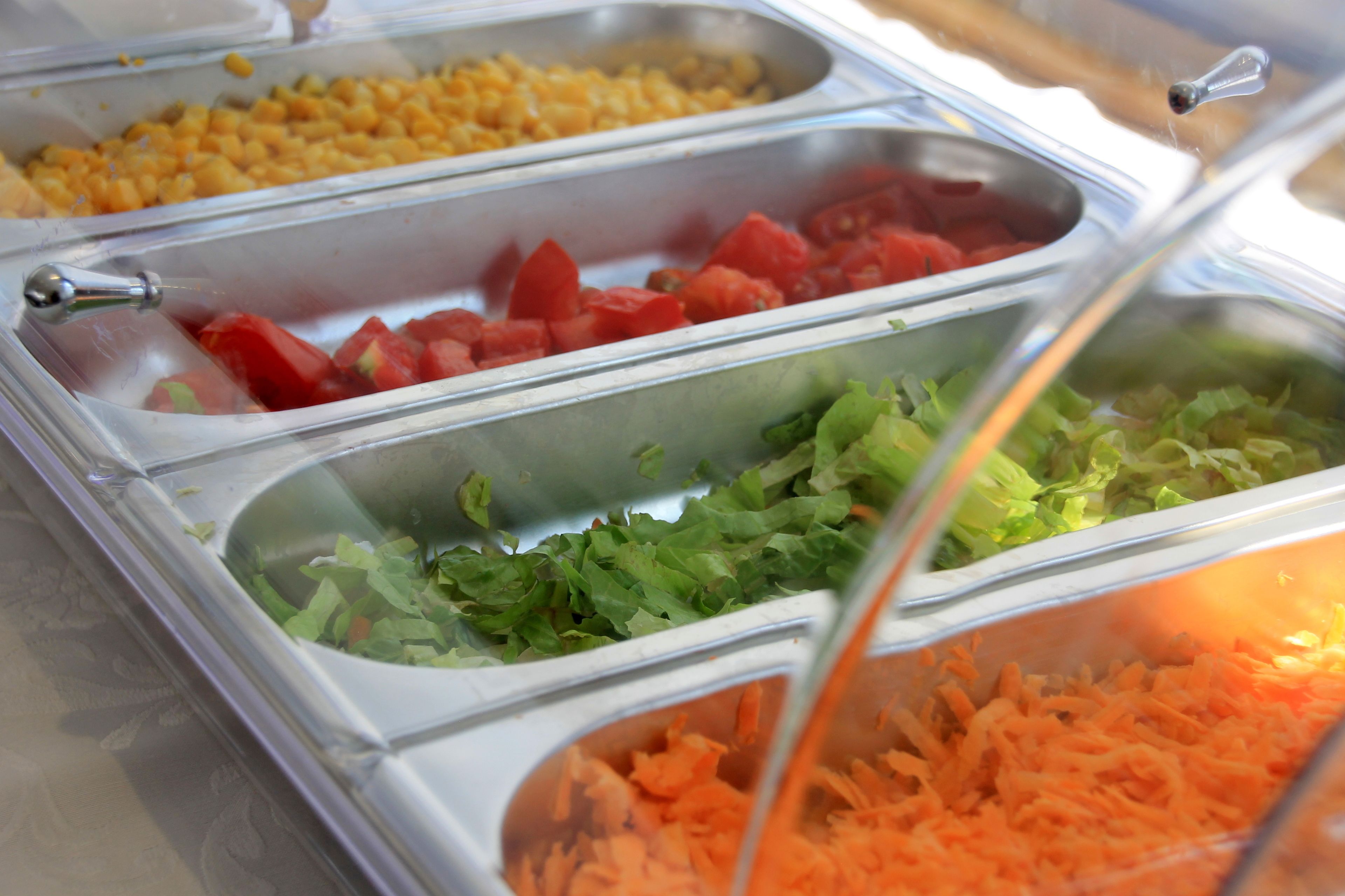 A buffet with various food items including corn, tomatoes, lettuce, and shredded carrots.