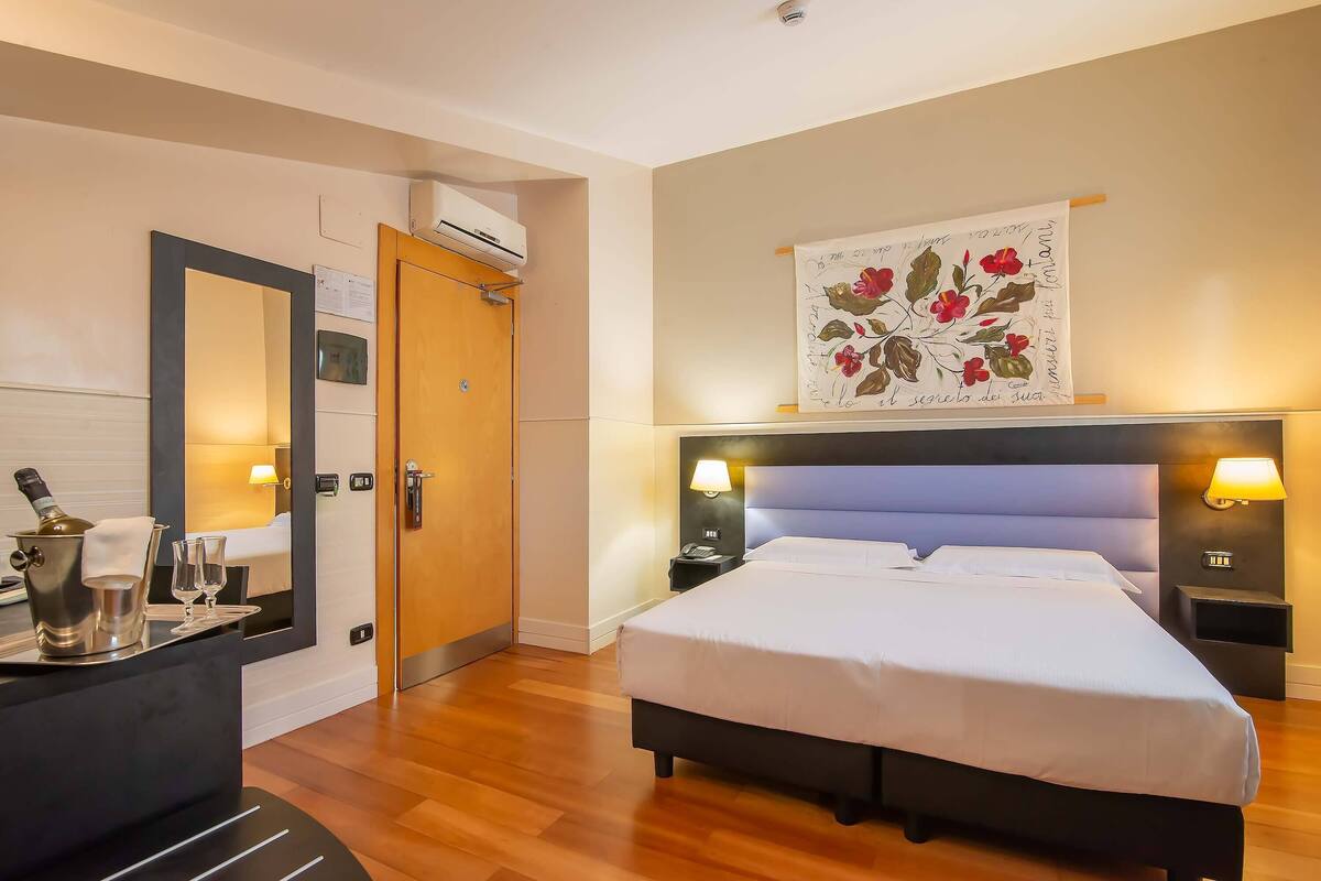Una camera d'albergo con un letto, abat-jour, comodini, una scrivania con una bottiglia e dei bicchieri, uno specchio e un'opera d'arte appesa al muro.