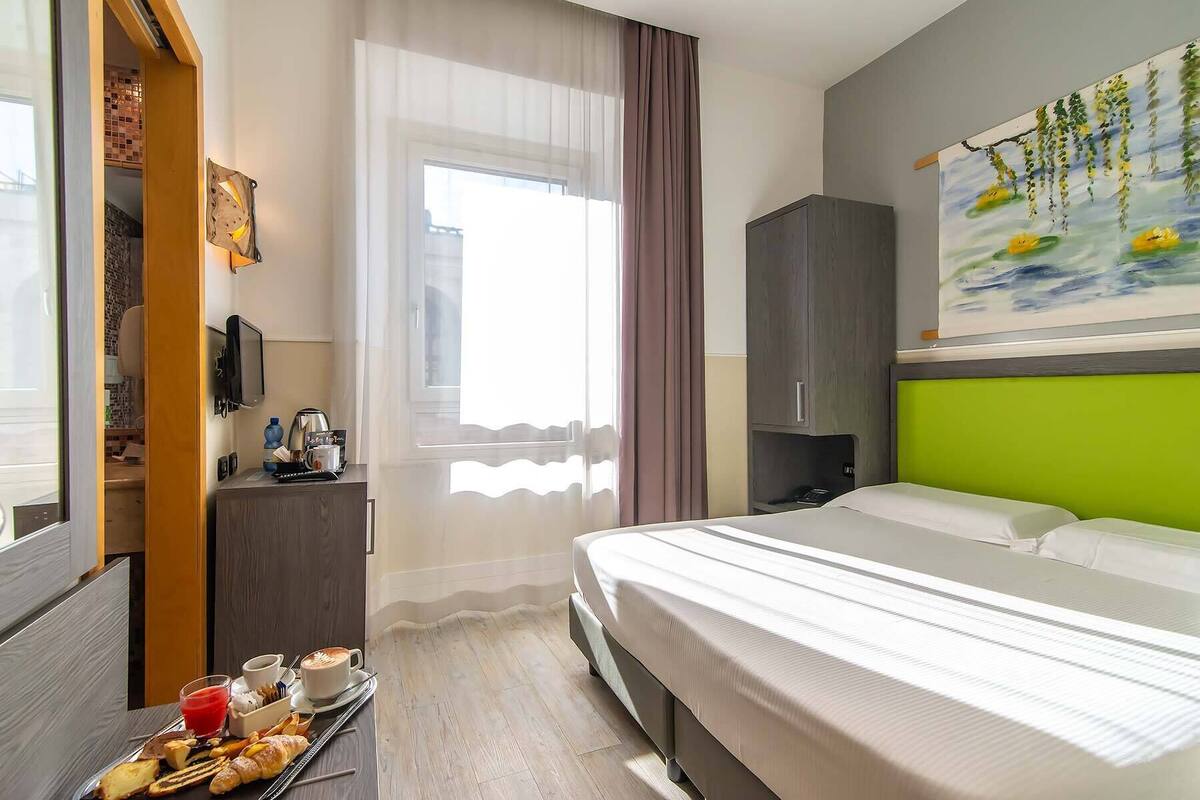 Una camera d'albergo con un letto, una scrivania con colazione inclusa, una televisione e un quadro appeso al muro.