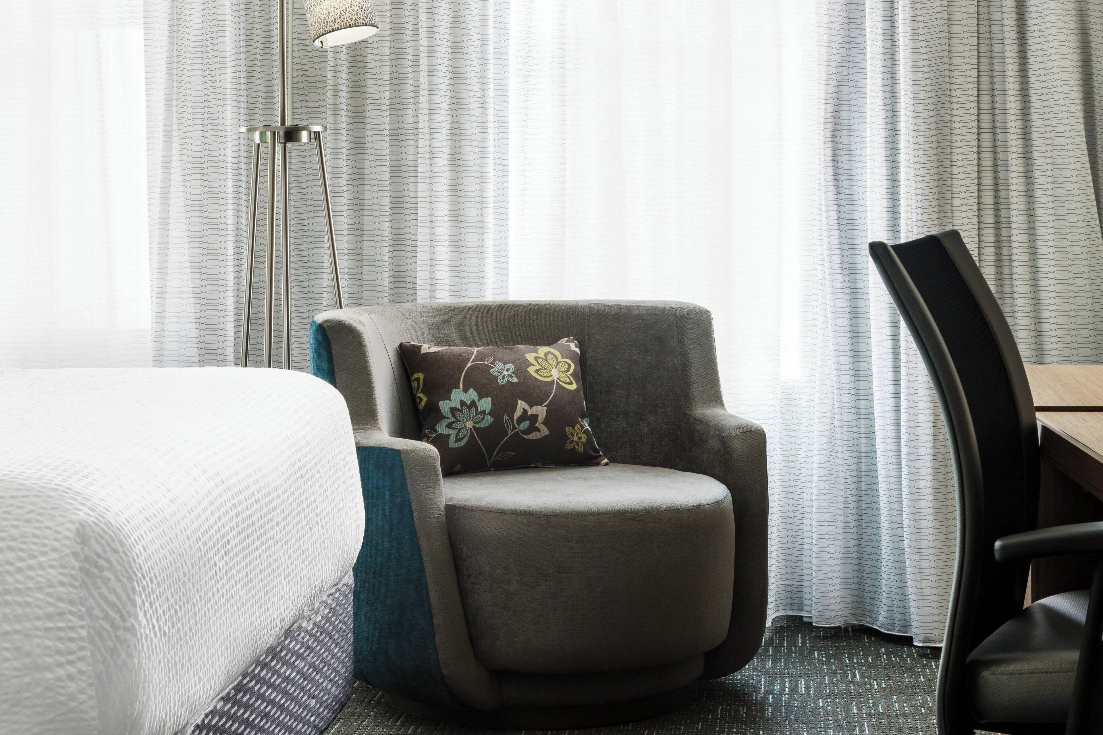 Une chambre d’hôtel avec un lit, un fauteuil gris orné d’un coussin à motifs floraux, un lampadaire et un bureau en bois.