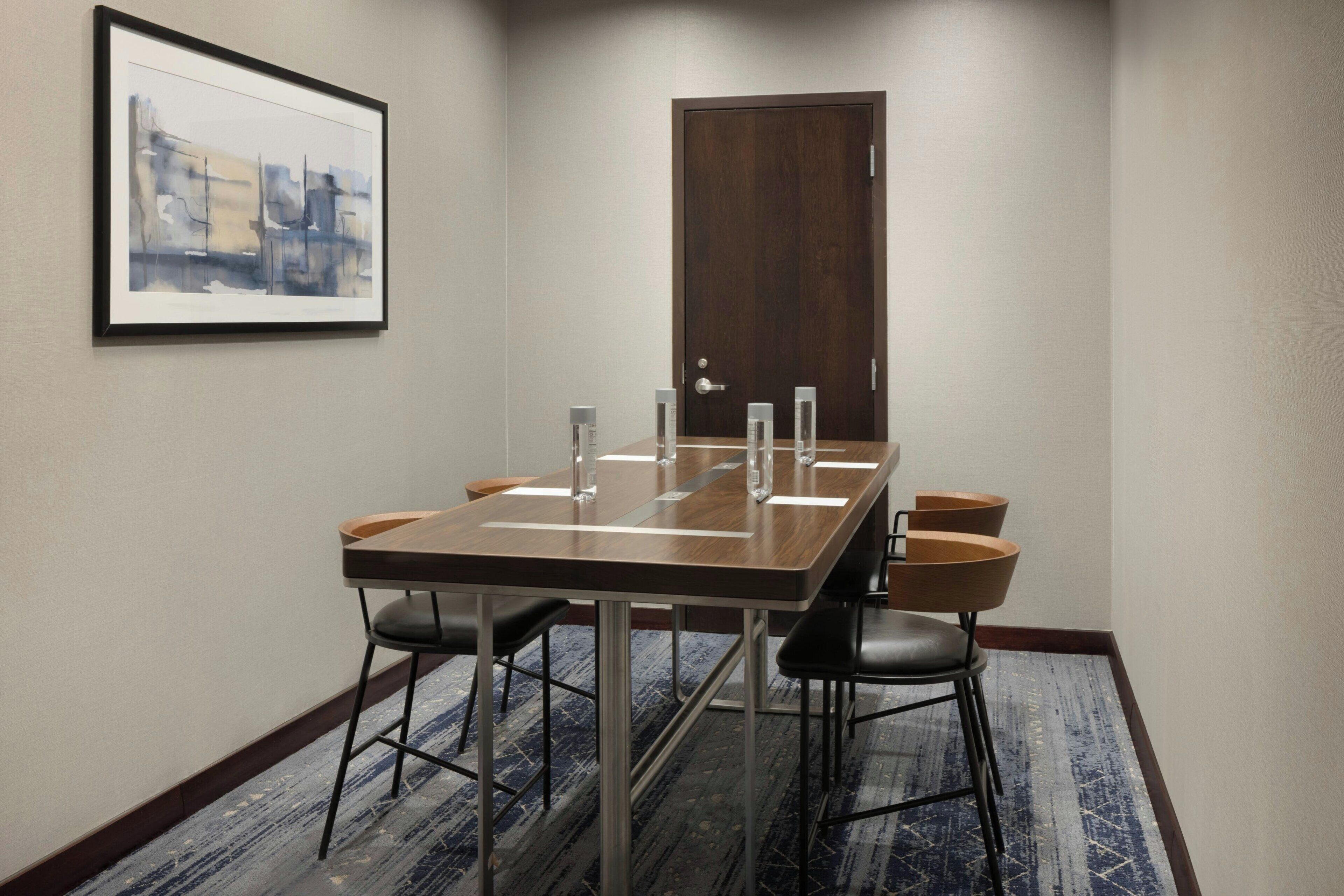 A conference room with a long table, four chairs, a framed abstract painting, a wooden door, and a patterned carpet.