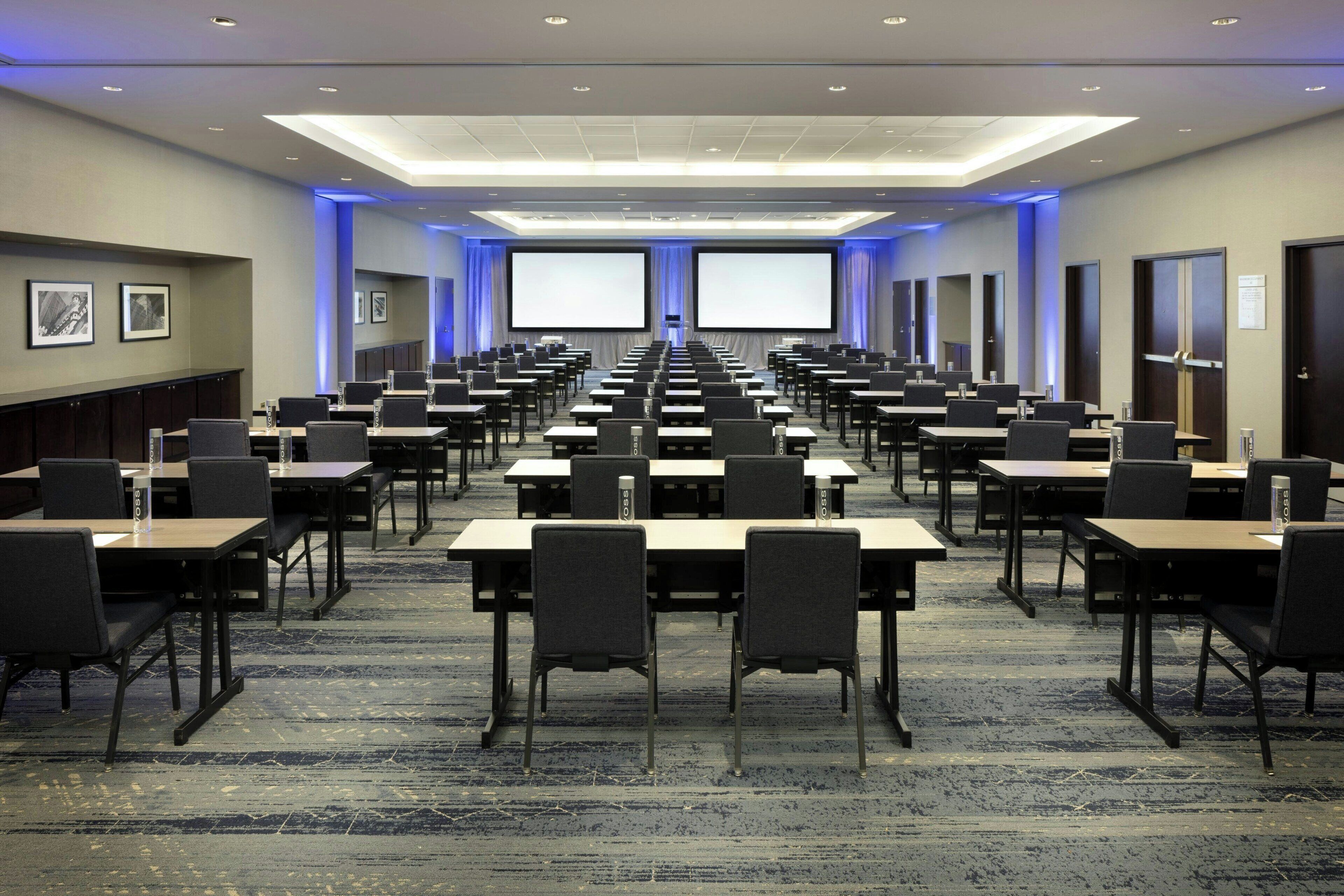 Un salón de conferencias con filas de mesas y sillas, pantallas grandes y piso alfombrado.