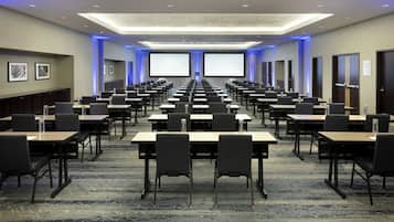 Un salón de conferencias con filas de mesas y sillas, pantallas grandes y piso alfombrado.