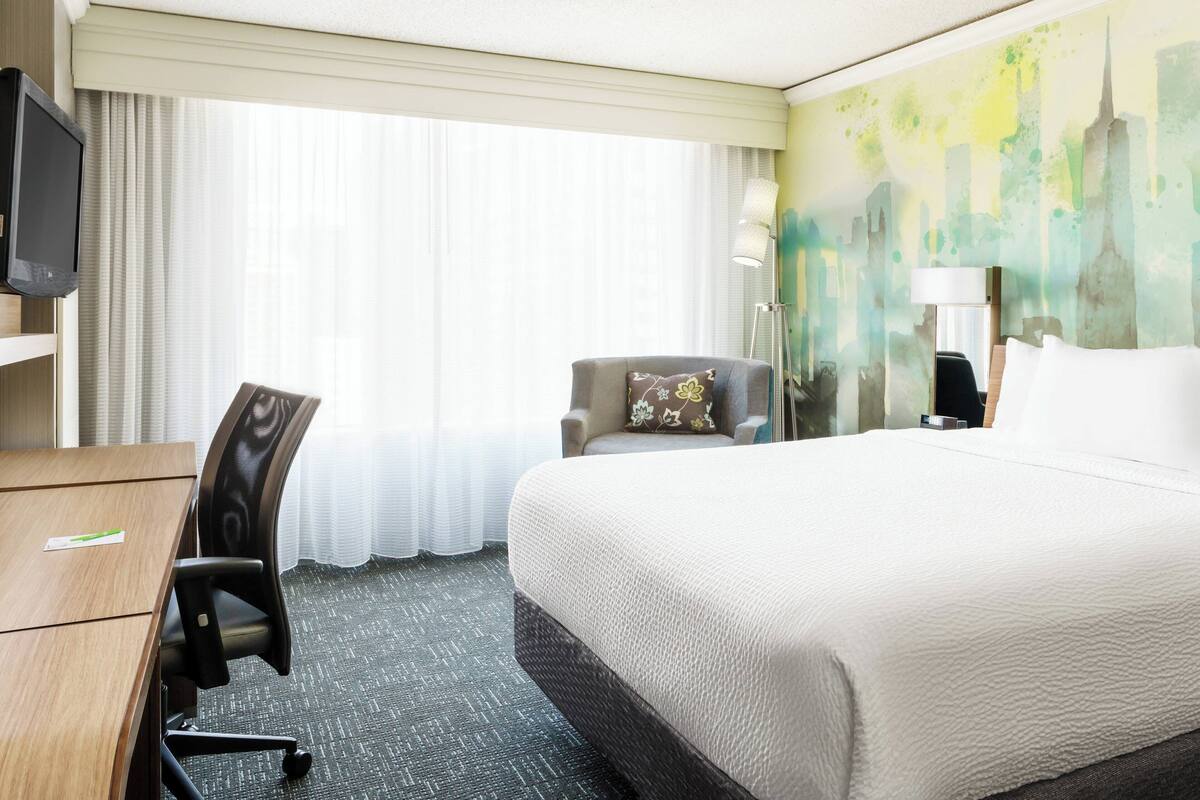 A hotel room with a bed, a desk, a chair, a TV, and a window with curtains.