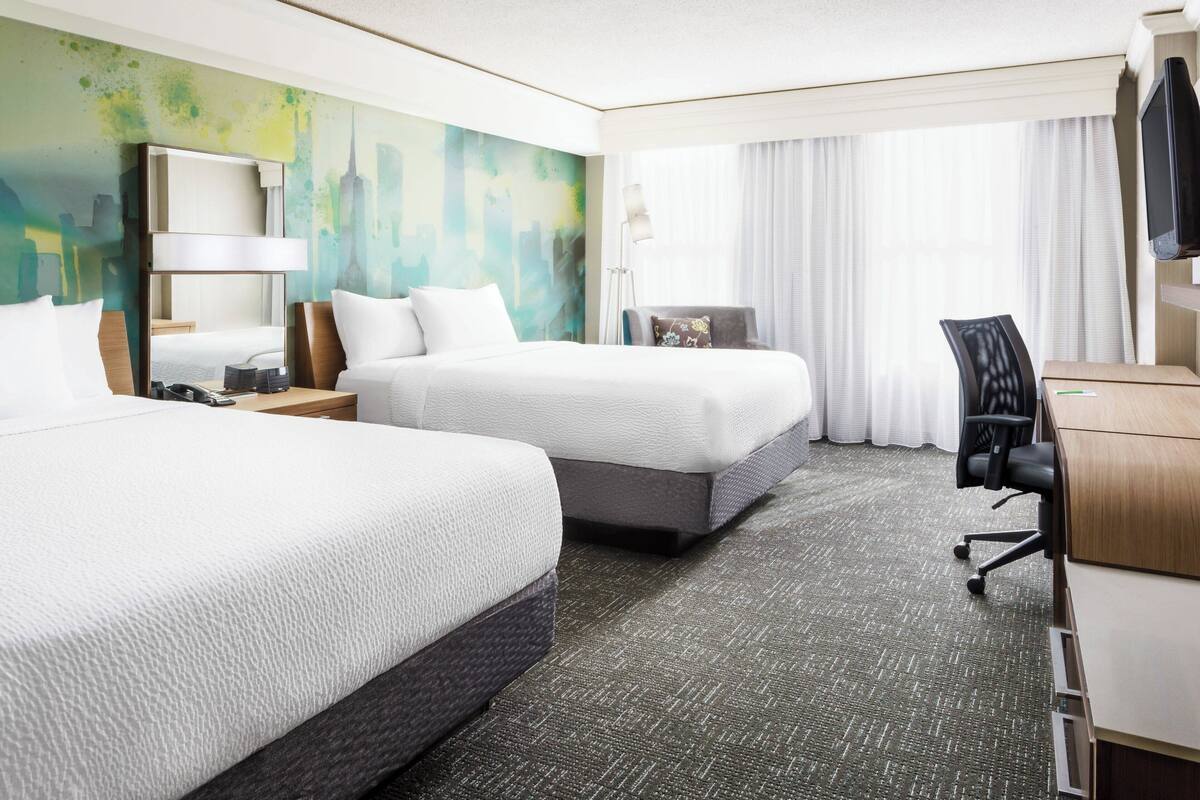 A hotel room with two beds, a desk, a chair, a lamp, and a large window with curtains.