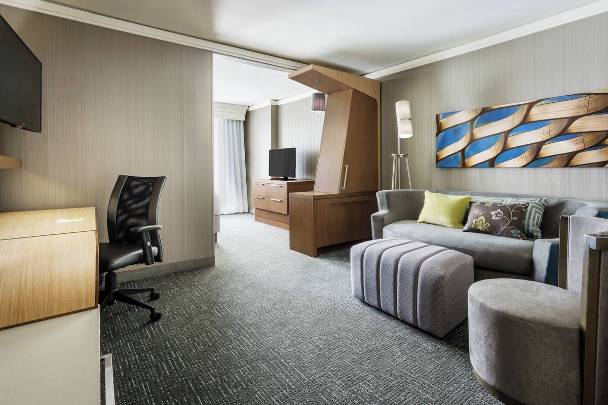 A modern hotel room with a desk, office chair, sofa, ottoman, and a television.