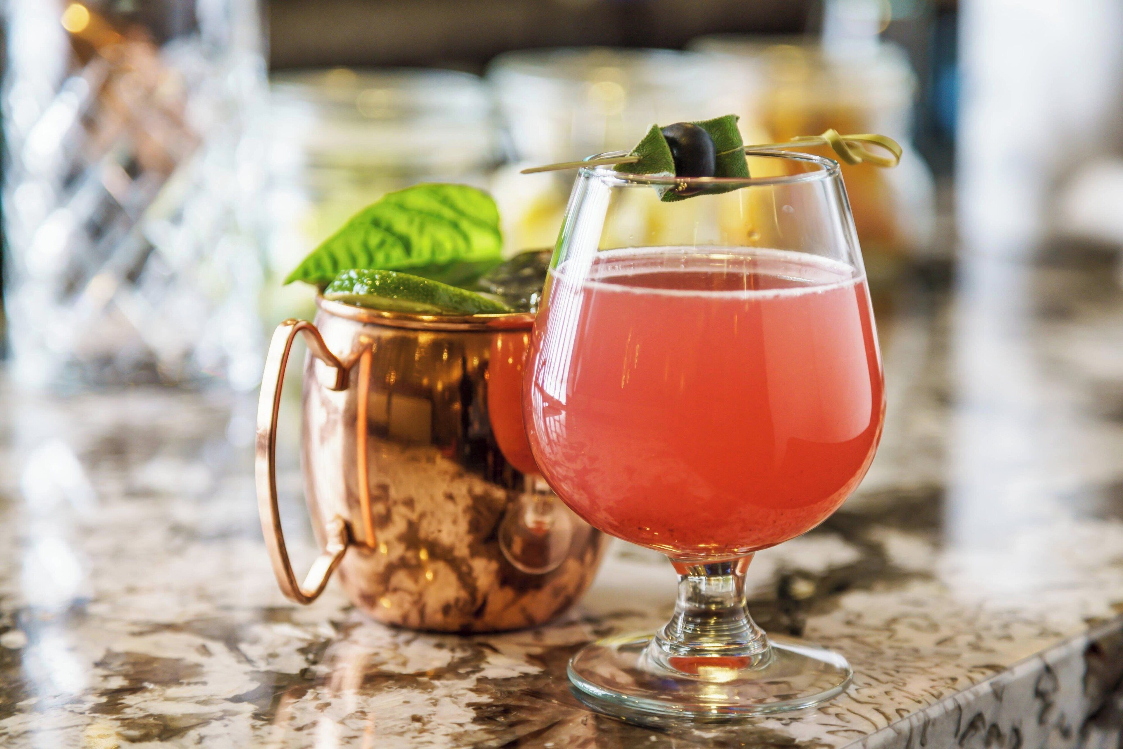A copper mug and a glass with a red drink, garnished with a leaf, on a marble surface.