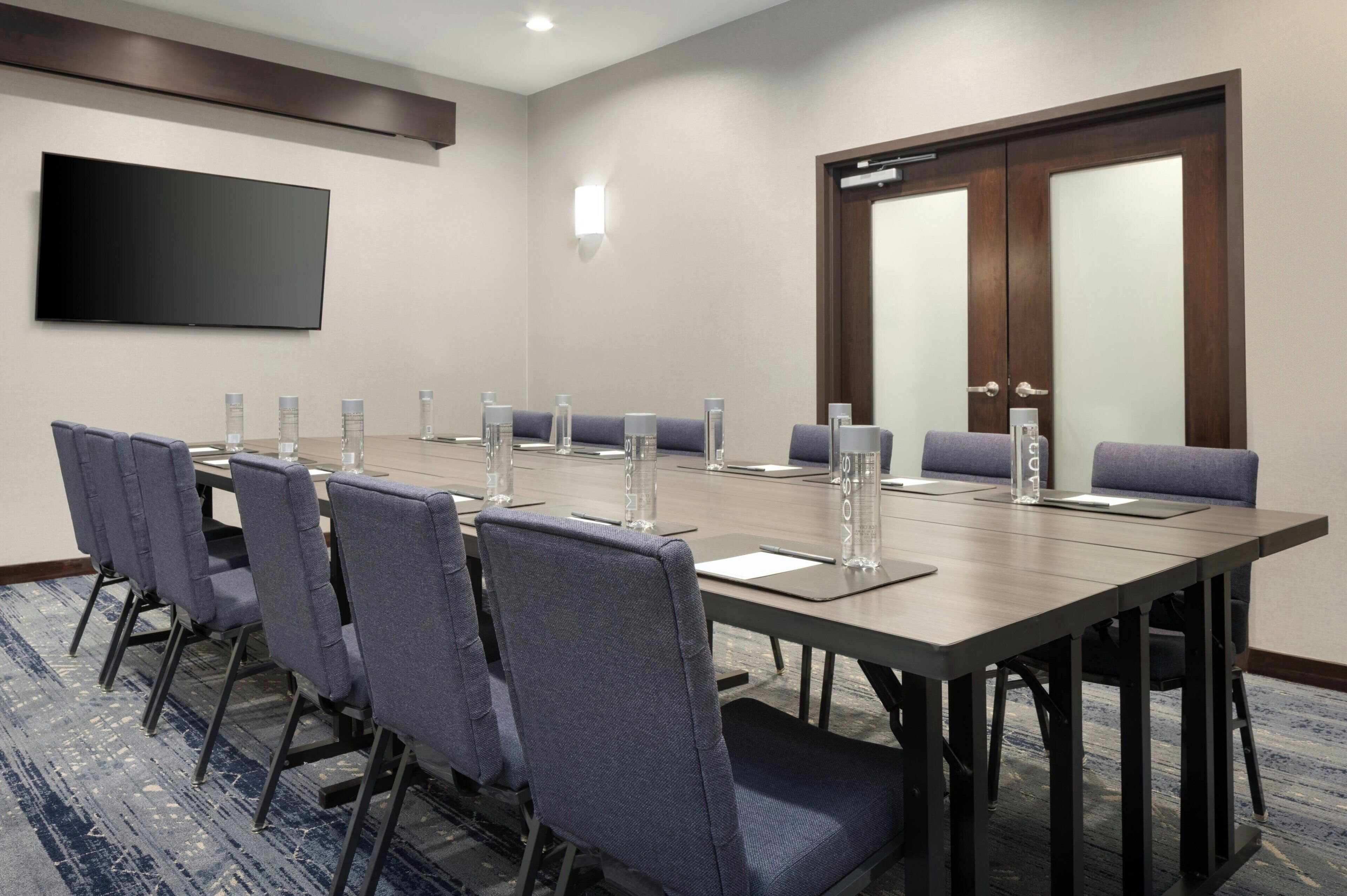 Una sala de conferencias con una larga mesa, sillas, un televisor de pantalla plana y una puerta con paneles de vidrio esmerilado.