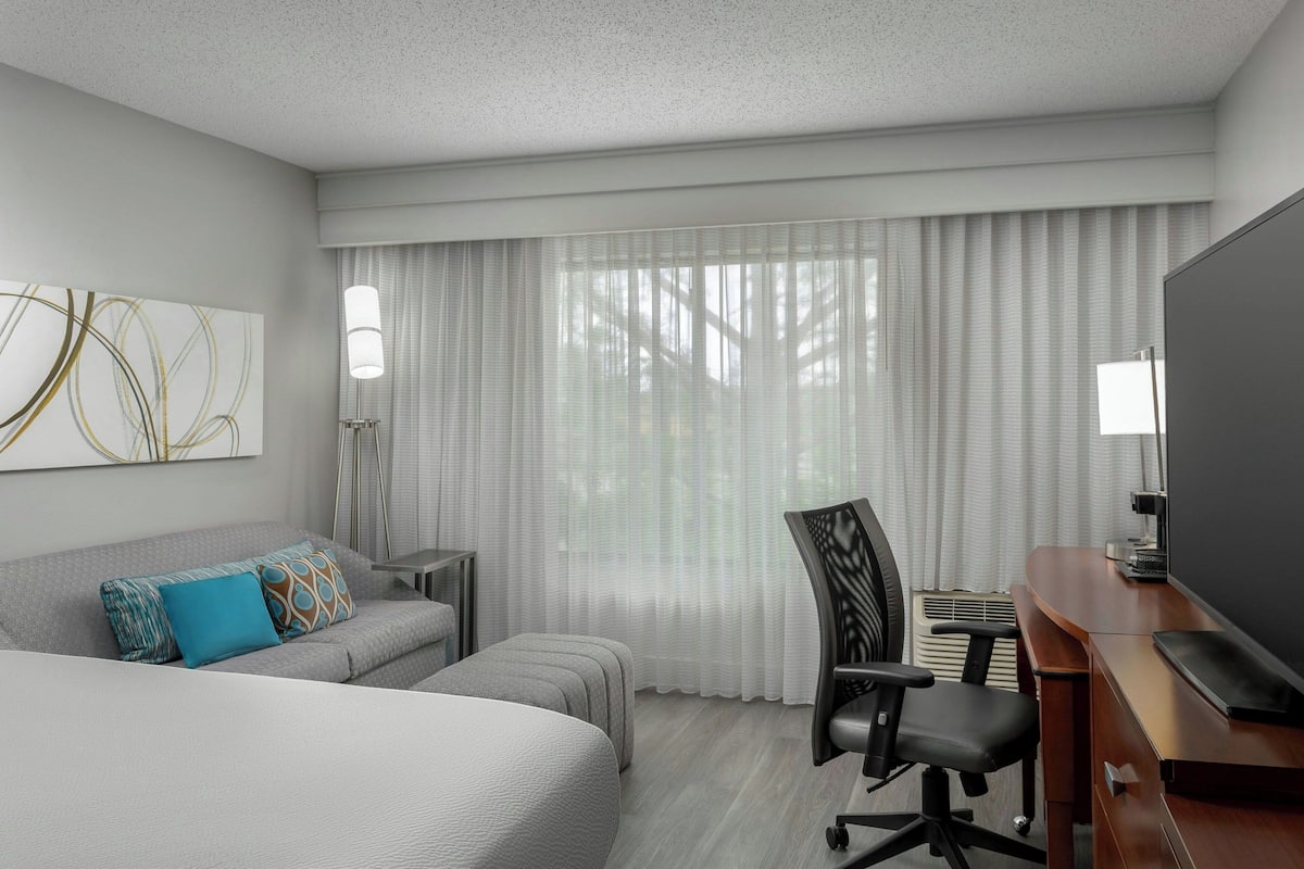 A hotel room with a large bed, a desk with a flat-screen TV, a chair, a lamp, and a window with sheer curtains.