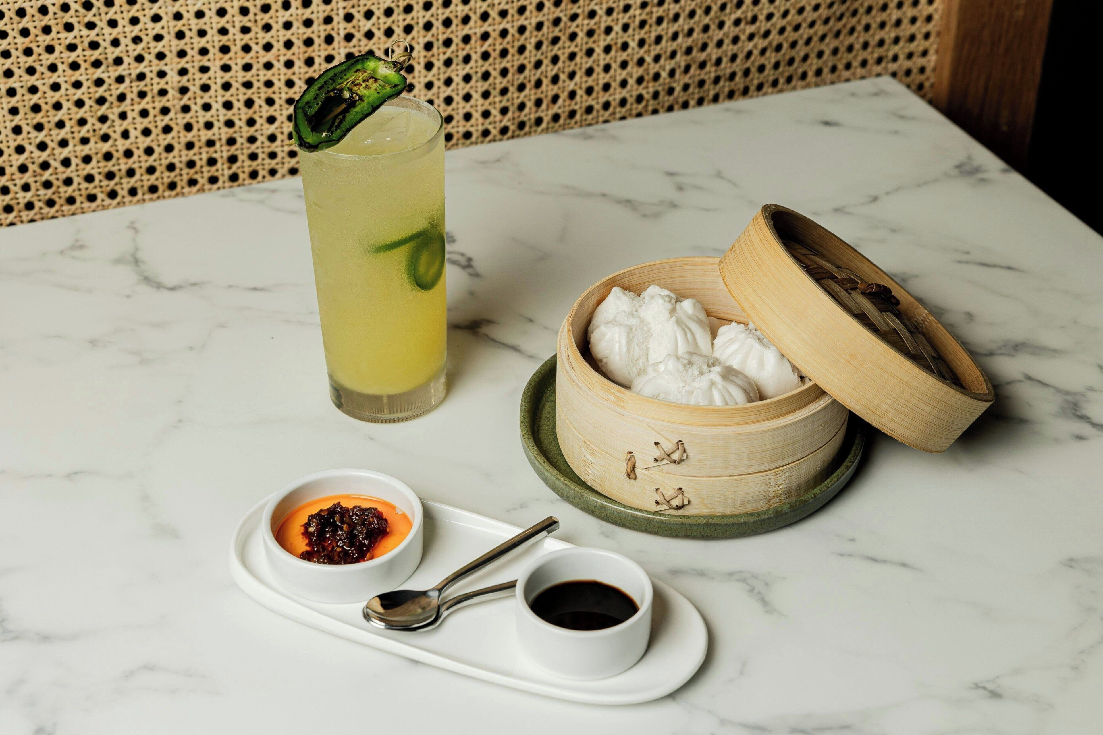 A table with a drink, a bamboo container with a lid, a small bowl with sauce, and a spoon on a tray.