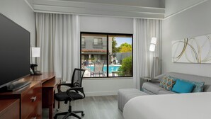 A hotel room with a bed, a desk with a computer, a chair, a TV, and a window with a view of a pool and buildings. - Courtyard by Marriott Houston Rice University (Houston)