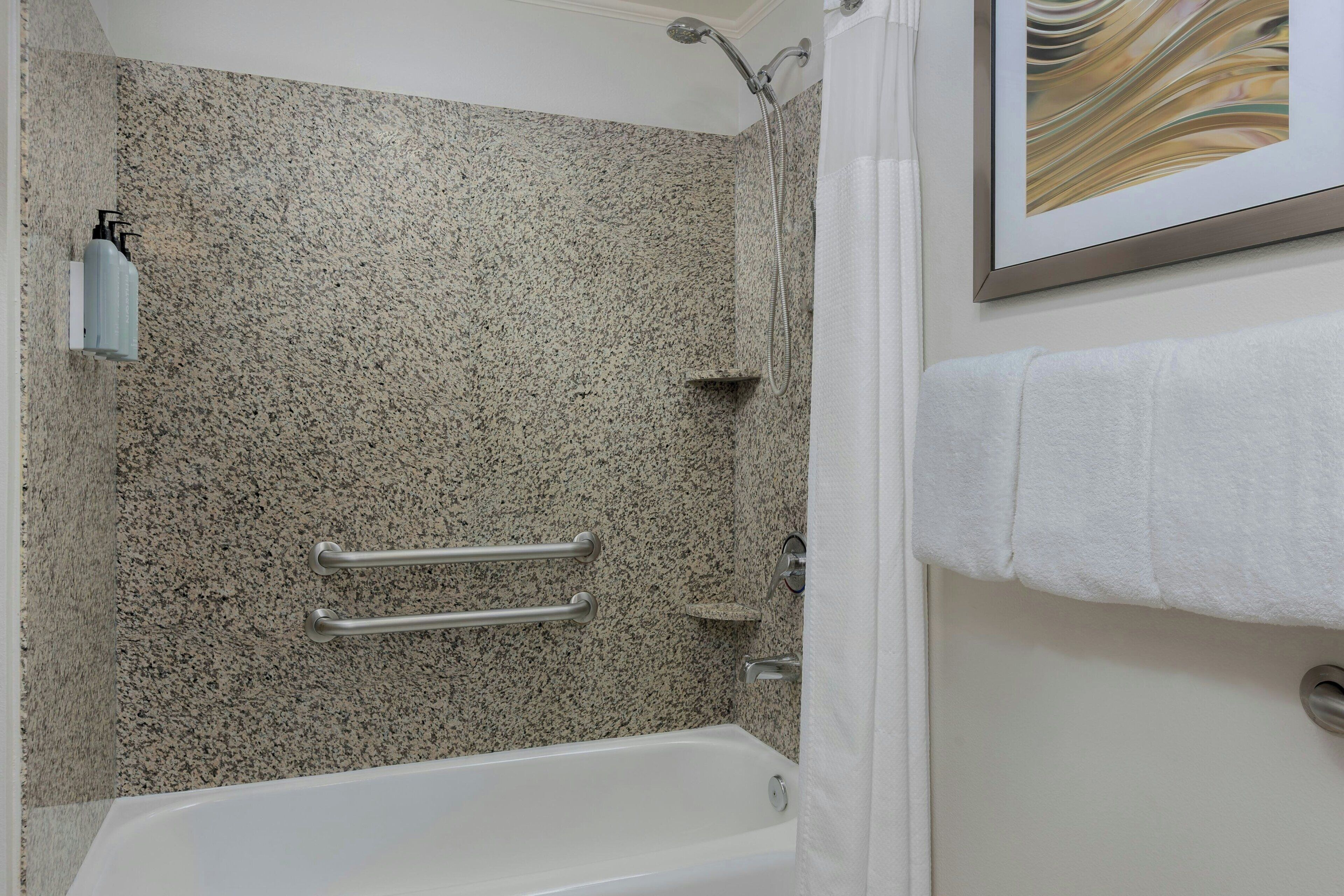 A bathroom with a granite shower wall, a white bathtub, and a framed artwork above the bathtub.