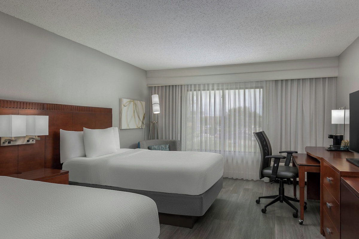 A hotel room with two beds, a desk, a chair, and a large window with curtains.