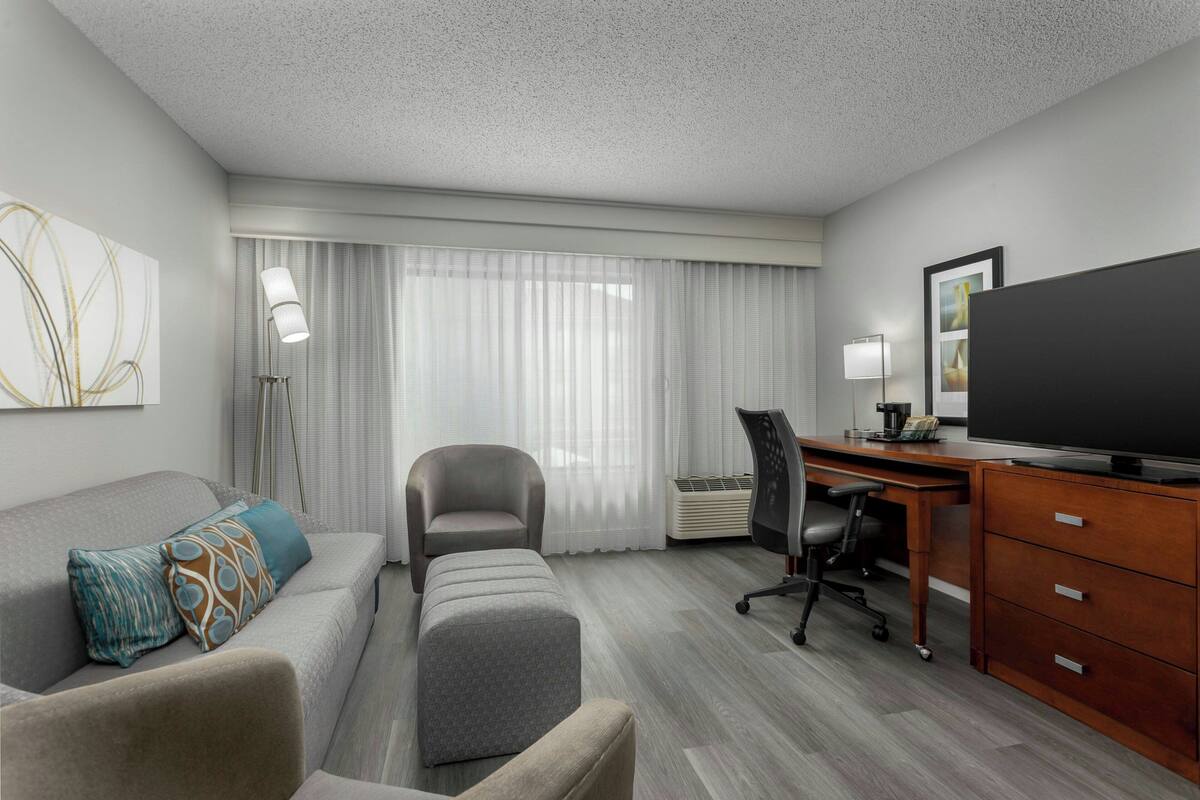 A hotel room with a grey sofa, a grey ottoman, a desk with a chair, a flat-screen TV, and a large window with sheer curtains.