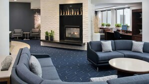A modern living room with a fireplace, grey sofas, a coffee table, and a desk area with plants. - Courtyard by Marriott Ewing Princeton (Trenton)