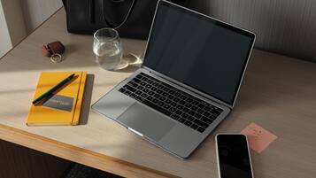 Un ordinateur portable est posé sur une table en bois, accompagné d’un carnet, d’un stylo, d’un verre d’eau, d’un téléphone intelligent et d’un petit mot.