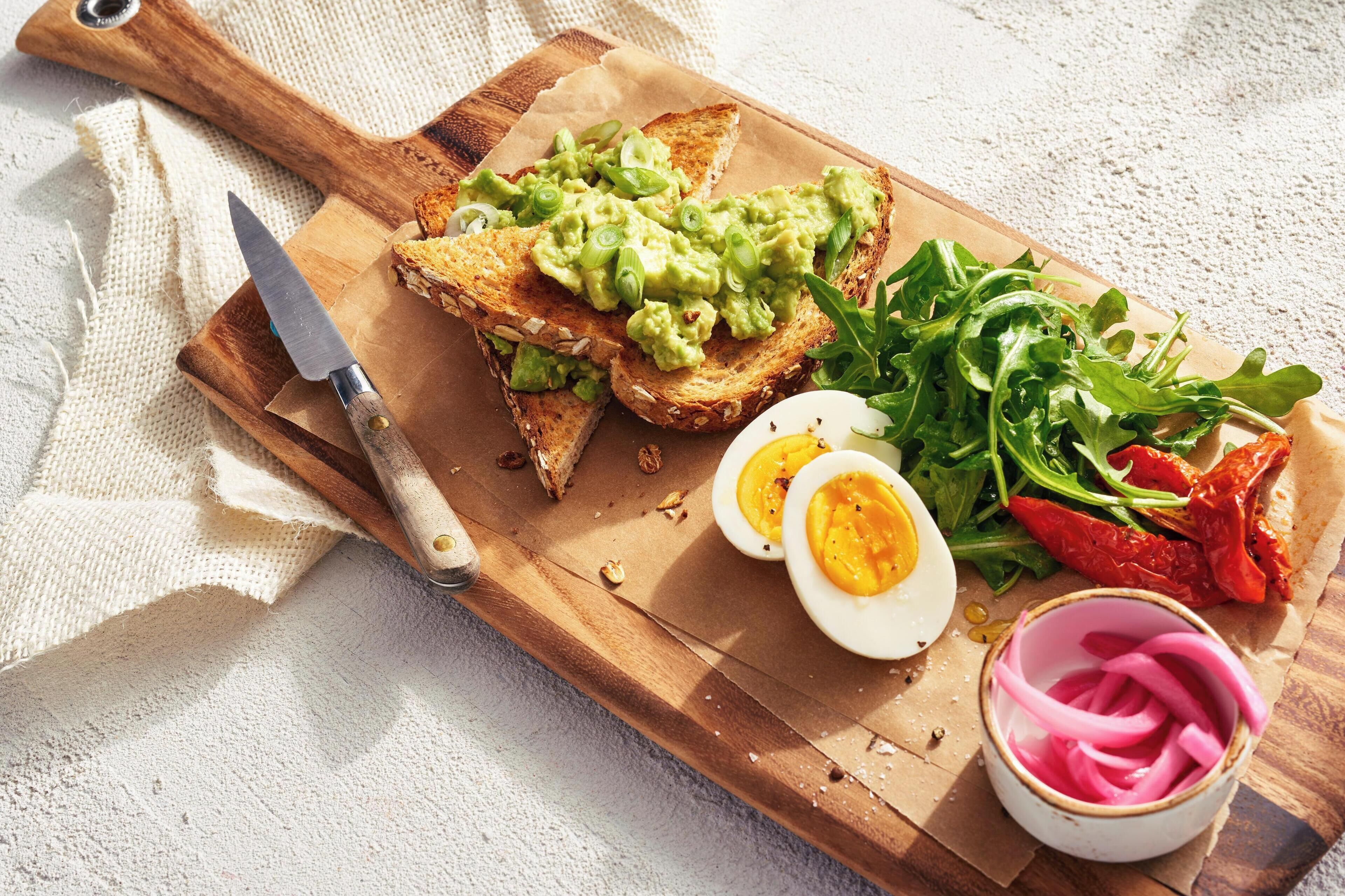 A wooden cutting board with toast, avocado, eggs, greens, and pickles.